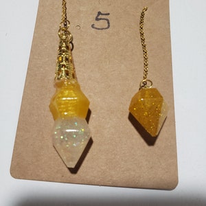 May include: Two gold-tone chain pendulums with yellow and white resin tips. The larger pendulum has a gold-tone metal top. The smaller pendulum has a yellow resin tip with gold glitter. The pendulums are displayed on a brown card with the number "5" written on it.