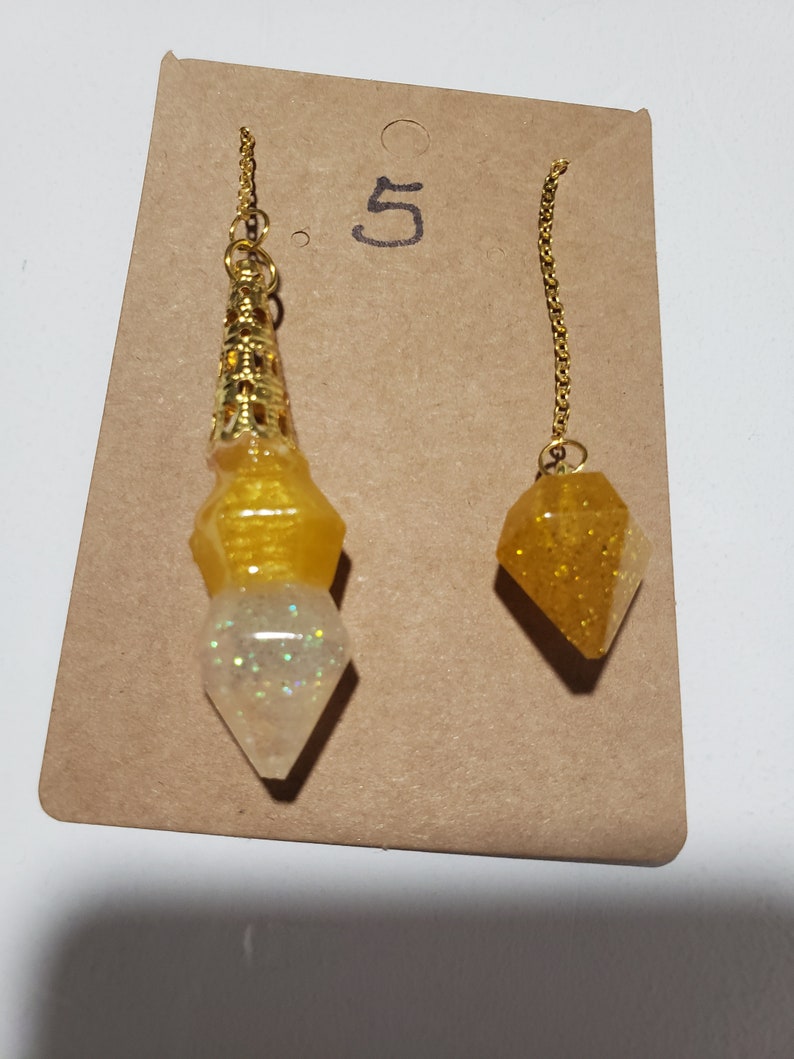 May include: Two gold-tone chain pendulums with yellow and white resin tips. The larger pendulum has a gold-tone metal top. The smaller pendulum has a yellow resin tip with gold glitter. The pendulums are displayed on a brown card with the number "5" written on it.