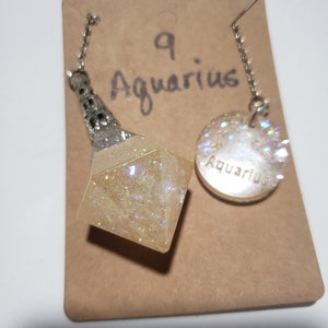May include: A white and silver necklace with a clear, sparkly, geometric pendant and a round charm with the word "Aquarius" on it. The necklace is on a brown card with the number "9" and the word "Aquarius" written on it.