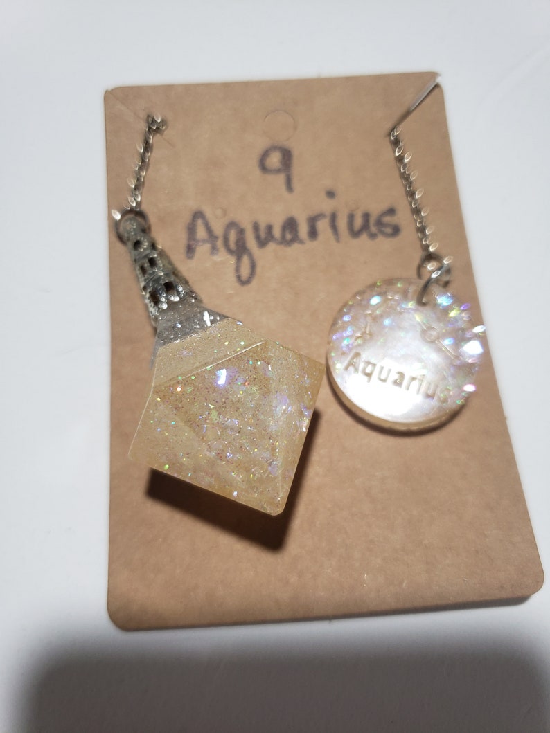May include: A white and silver necklace with a clear, sparkly, geometric pendant and a round charm with the word "Aquarius" on it. The necklace is on a brown card with the number "9" and the word "Aquarius" written on it.