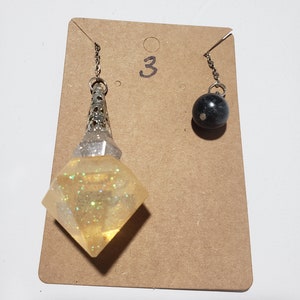 May include: Two silver chain pendulums with different charms. One charm is a clear, diamond-shaped crystal with silver glitter. The other charm is a black sphere. The pendulums are hanging from a brown card with the number "3" written on it.