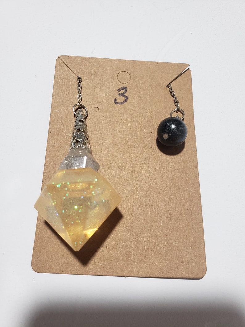 May include: Two silver chain pendulums with different charms. One charm is a clear, diamond-shaped crystal with silver glitter. The other charm is a black sphere. The pendulums are hanging from a brown card with the number "3" written on it.