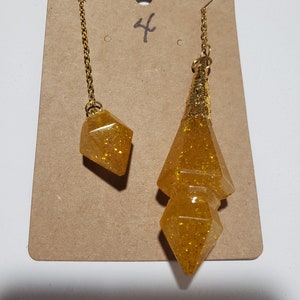 May include: Two gold-toned pendulums with a glitter finish. The larger pendulum is a double-pointed teardrop shape. The smaller pendulum is a diamond shape. Both pendulums are hanging from gold chains.