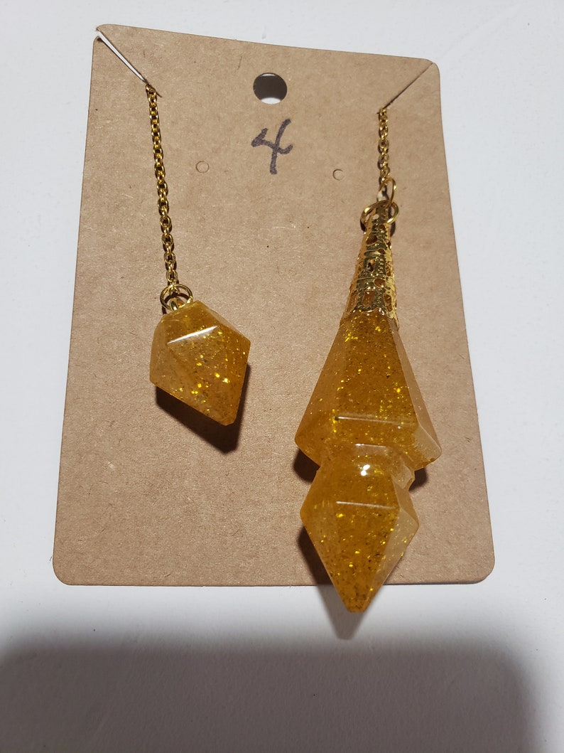 May include: Two gold-toned pendulums with a glitter finish. The larger pendulum is a double-pointed teardrop shape. The smaller pendulum is a diamond shape. Both pendulums are hanging from gold chains.