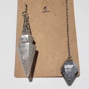 May include: Two silver and grey crystal pendulums with chains hanging from a brown card with the number "1" written on it.