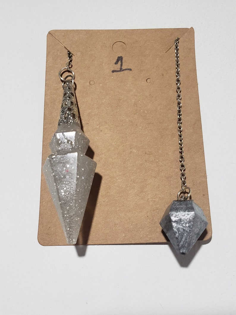 May include: Two silver and grey crystal pendulums with chains hanging from a brown card with the number "1" written on it.