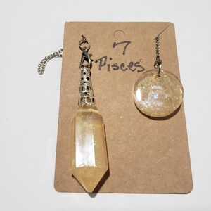 May include: A set of two gold-toned, glitter-filled, resin pendants on chains. The larger pendant is round and the smaller pendant is a pointed, crystal-shaped charm. The pendants are labelled "7 Pisces" on a brown card.