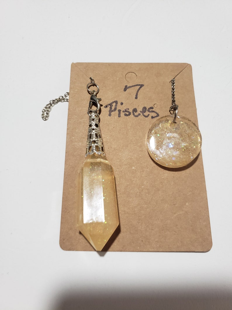 May include: A set of two gold-toned, glitter-filled, resin pendants on chains. The larger pendant is round and the smaller pendant is a pointed, crystal-shaped charm. The pendants are labelled "7 Pisces" on a brown card.