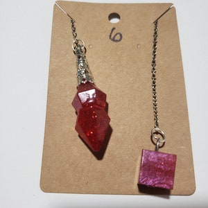 May include: Two red crystal pendulums on a brown card with the number "6" written on it. One pendulum is a pointed shape and the other is a square shape.