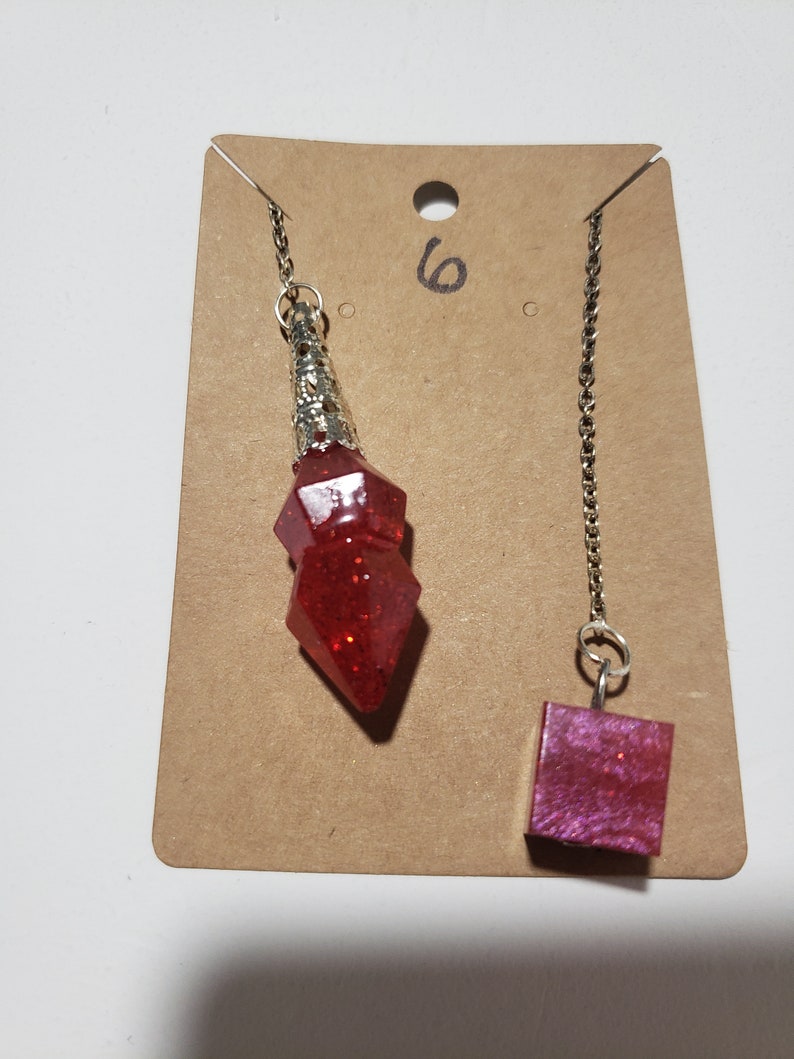 May include: Two red crystal pendulums on a brown card with the number "6" written on it. One pendulum is a pointed shape and the other is a square shape.