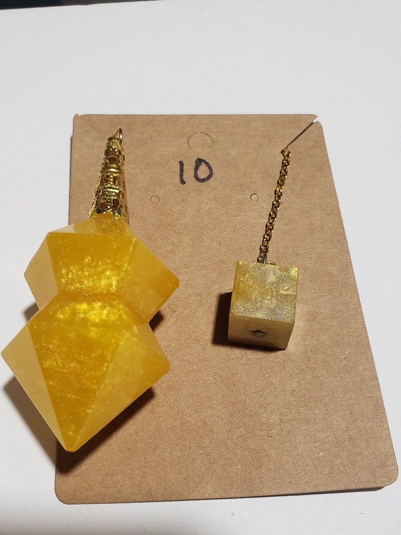 May include: A gold-coloured, double-pointed, crystal pendulum with a gold chain and a small, gold-coloured cube. The pendulum is on a brown card with the number "10" written on it.