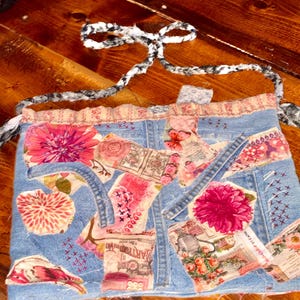 Könnte beinhalten: Eine handgefertigte Jeansschürze mit Blumen- und Vintage-Papier-Patchwork. Die Schürze hat eine hellblaue Jeansbasis, verziert mit rosa und roten Blumenmustern und einem schwarz-weiß gemusterten Bindeband.