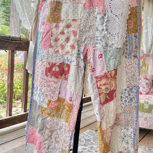 Könnte beinhalten: Patchworkhose mit einer Vielzahl von Blumen- und gemusterten Stoffen in Rosa, Weiß, Blau und Gelb. Die Hose hat einen blauen Bund und weiße Spitzendetails. Grün-weiß karierte Bänder sind an den Knöcheln gebunden.