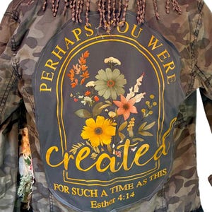 May include: A camouflage jacket featuring a dark gray back panel with a floral graphic and the text "PERHAPS YOU WERE created FOR SUCH A TIME AS THIS Esther 4:14". The jacket includes brown crocheted fringe at the collar and black fringe at the hem.