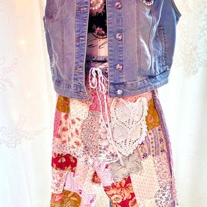 May include: A denim vest with jewel buttons and a colourful crocheted patch is layered over a floral top and patchwork trousers. The trousers feature a mix of floral and patterned fabrics in shades of pink, red, and cream. A white lace detail is on the trousers.