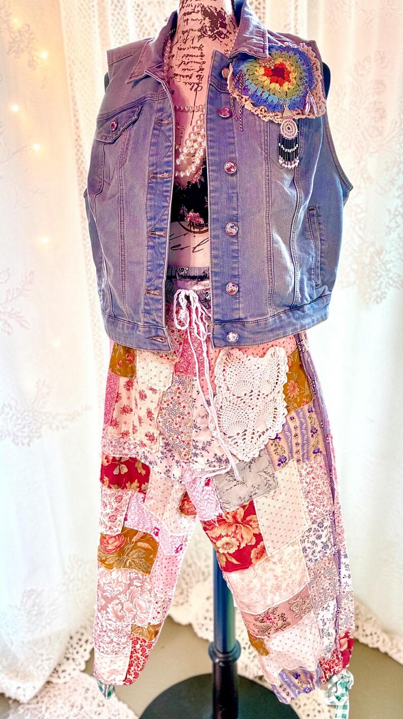 May include: A denim vest with jewel buttons and a colourful crocheted patch is layered over a floral top and patchwork trousers. The trousers feature a mix of floral and patterned fabrics in shades of pink, red, and cream. A white lace detail is on the trousers.