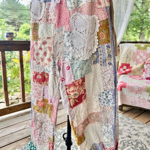 May include: Patchwork trousers in a variety of floral and patterned fabrics, including pink, blue, and white. The trousers have a drawstring waist and are decorated with white lace doilies. The cuffs are gathered with green and white checkered fabric.