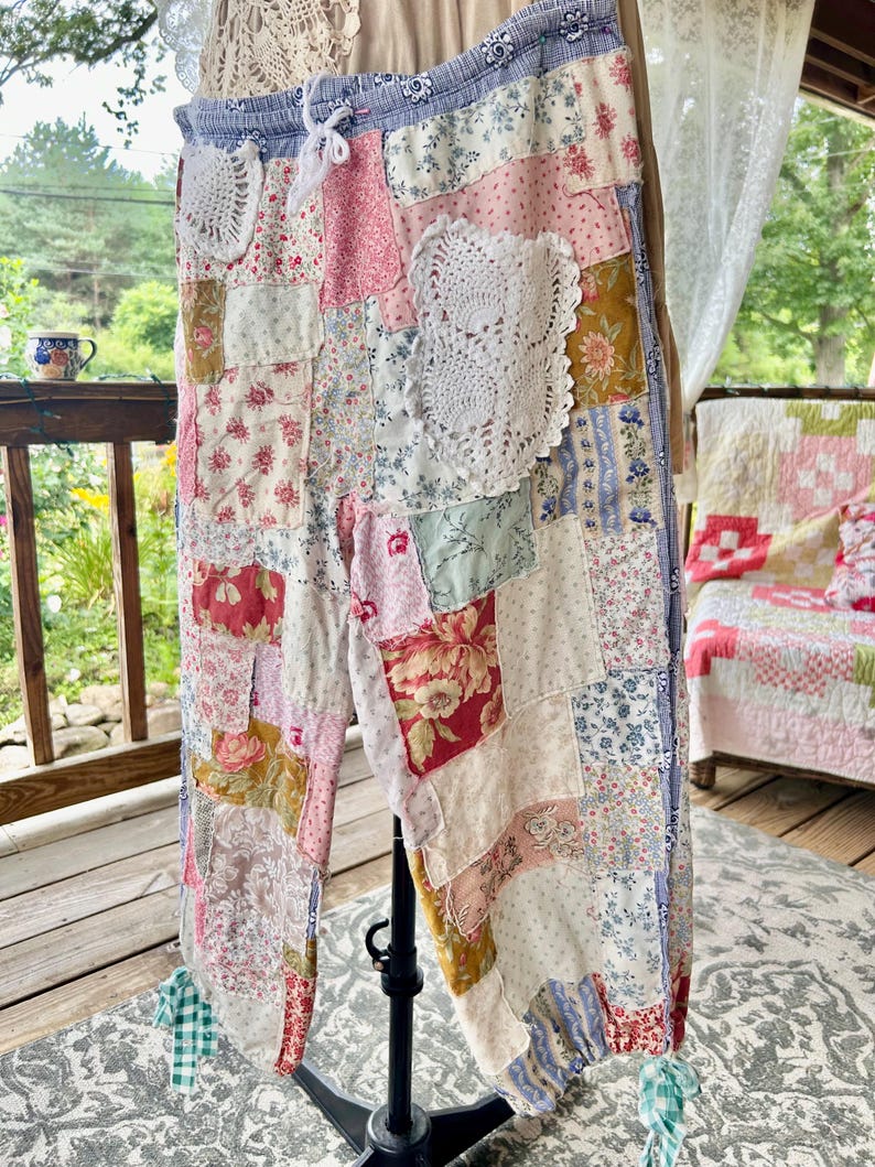 May include: Patchwork trousers in a variety of floral and patterned fabrics, including pink, blue, and white. The trousers have a drawstring waist and are decorated with white lace doilies. The cuffs are gathered with green and white checkered fabric.