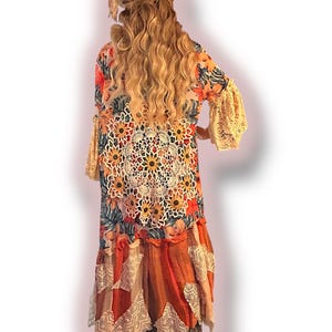 May include: A long, flowing, floral print maxi dress with a crocheted overlay and lace trim. The dress has a loose, bohemian style and features a large crocheted mandala design on the front. The dress is paired with a straw hat.