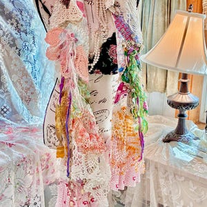 May include: A multi-layered, bohemian-style scarf on a mannequin. The scarf features various textures, including crochet, lace, and fabric scraps in shades of white, pink, orange, and green. Embellishments include beads and ribbons, creating a unique, handcrafted accessory.