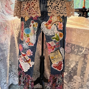 May include: Blue denim jeans with floral and lace patches, adorned with fabric flowers and a ruffled hem. A light blue floral jacket with lace trim is draped over the jeans. The jeans are displayed on a black stand.