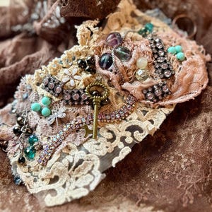 May include: A delicate, handmade bracelet featuring a mix of lace, beads, and a vintage key charm. The bracelet is adorned with turquoise, pink, and clear crystals, creating a whimsical and romantic look.