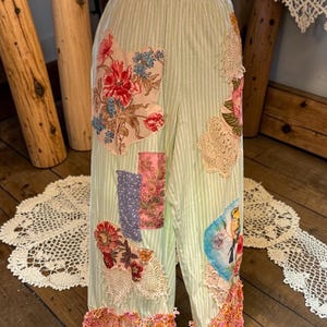 May include: Light green striped pants with floral patchwork and lace accents. The pants feature a high waist and a wide leg with a pink, fringed hem. Patches include floral and patterned fabrics. The pants are a unique, bohemian style.