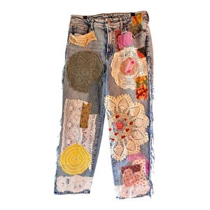 Puede incluir: Vaqueros de mezclilla de lavado claro con varios parches de tela y tapetes de encaje. Los parches incluyen telas florales, de cuadros y lisas en tonos rosa, amarillo, verde y marrón. Los vaqueros tienen bordes deshilachados y un aspecto único y artesanal.