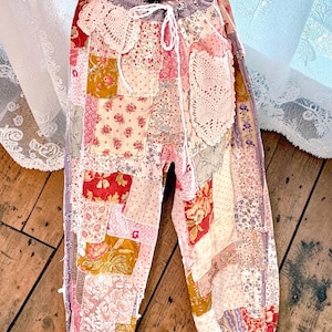 May include: Patchwork trousers with a drawstring waist. The trousers feature a variety of floral and patterned fabrics in shades of pink, red, yellow, and white. The design includes lace accents and gathered cuffs, creating a unique, bohemian style.