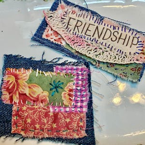 May include: Two rectangular fabric patches featuring frayed denim edges and layered textile scraps. One patch displays the word FRIENDSHIP embroidered in black thread over a cream lace strip. The other features floral and checkered patterns.