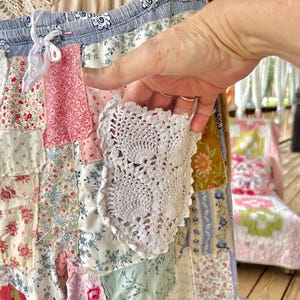 May include: A patchwork skirt featuring floral patterns in pink, red, blue, and yellow. The skirt has a blue and white patterned waistband with a drawstring. A white crocheted pocket is attached to the skirt.