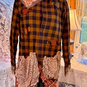 May include: Brown and navy plaid shirt with patches and crochet pockets. The shirt has a high collar and long sleeves with lace cuffs. Patches of fabric and lace are sewn onto the shirt, giving it a unique, handcrafted look.