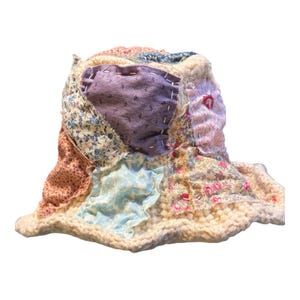 May include: A patchwork bucket hat featuring a variety of fabric patterns and colors, including floral prints, solid lavender, and light blue. The hat is constructed with visible stitching and a cream-colored, textured brim. The hat's design is a mix of vintage and modern styles.