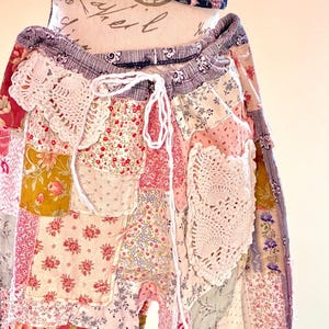 May include: Patchwork trousers in a mix of floral and patterned fabrics, predominantly pink, red, yellow, and blue. The trousers have a drawstring waist and crochet lace details. The top is a navy blue floral print.