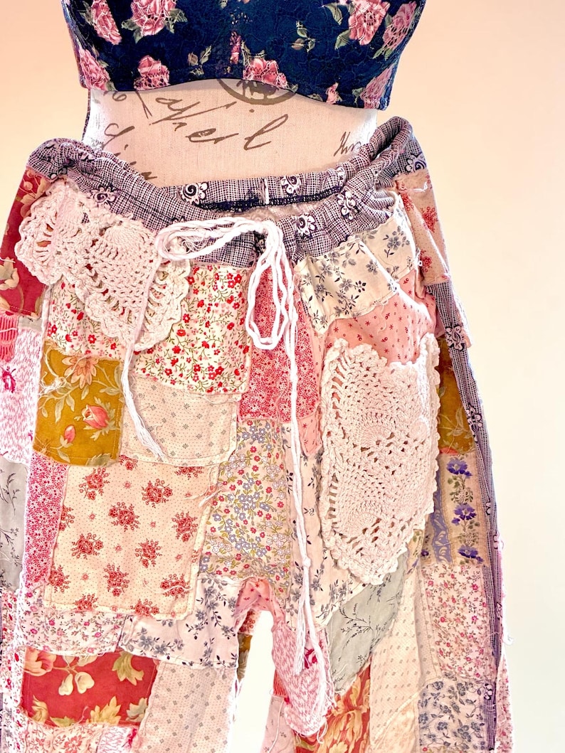 May include: Patchwork trousers in a mix of floral and patterned fabrics, predominantly pink, red, yellow, and blue. The trousers have a drawstring waist and crochet lace details. The top is a navy blue floral print.