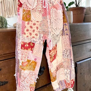 May include: Patchwork trousers in pink, red, and gold, with floral and patterned fabric squares. The trousers have a drawstring waist and are decorated with lace doilies and ribbons. The style is bohemian and vintage-inspired, with a relaxed fit.
