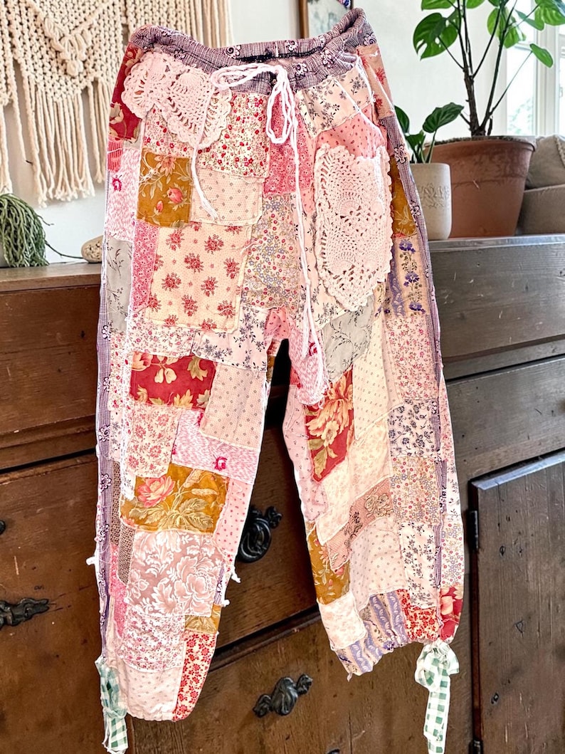 May include: Patchwork trousers in pink, red, and gold, with floral and patterned fabric squares. The trousers have a drawstring waist and are decorated with lace doilies and ribbons. The style is bohemian and vintage-inspired, with a relaxed fit.