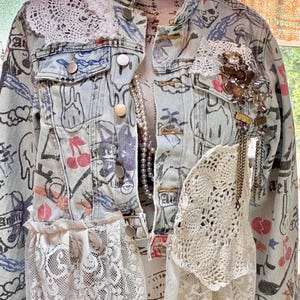 May include: Light blue denim jacket with a unique design. The jacket features hand-drawn graphics, lace accents, and decorative embellishments. It has a button-up front, pockets, and a collared neckline. A pearl necklace is visible beneath the jacket.