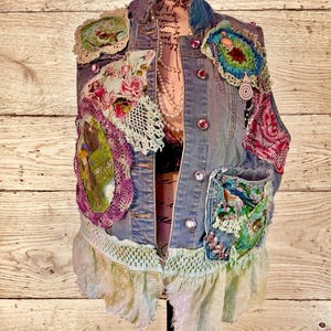 May include: A light blue denim vest adorned with colorful patches, lace, and floral appliques. The vest features a ruffled hem and decorative buttons. Patches include birds, flowers, and crochet details. The vest is displayed on a mannequin.