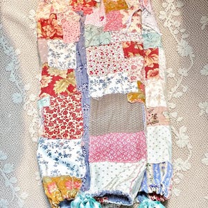 May include: A patchwork skirt in a mix of floral and patterned fabrics, including red, pink, blue, and cream. The skirt has a gathered waist and is finished with blue and white gingham bows. The design has a vintage, handmade aesthetic, with a variety of fabrics.
