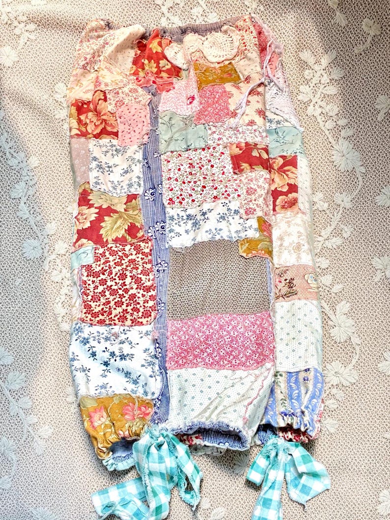 May include: A patchwork skirt in a mix of floral and patterned fabrics, including red, pink, blue, and cream. The skirt has a gathered waist and is finished with blue and white gingham bows. The design has a vintage, handmade aesthetic, with a variety of fabrics.