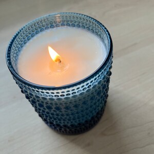 May include: A blue glass candle jar with a white wax candle and a lit wick. The jar has a textured surface.