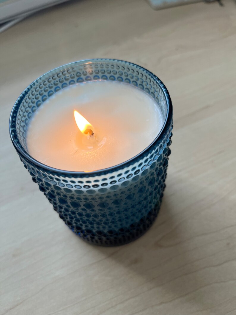 May include: A blue glass candle jar with a white wax candle and a lit wick. The jar has a textured surface.