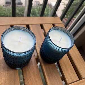 May include: Two blue glass candle jars with white wax and wicks, sitting on a wooden table.