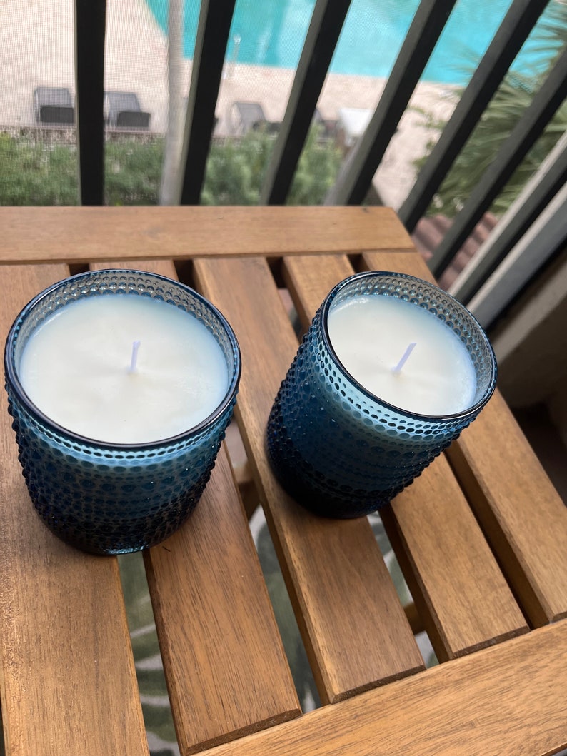 May include: Two blue glass candle jars with white wax and wicks, sitting on a wooden table.