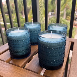 May include: Four blue glass candles with white wax and white wicks. The candles are sitting on a wooden table.
