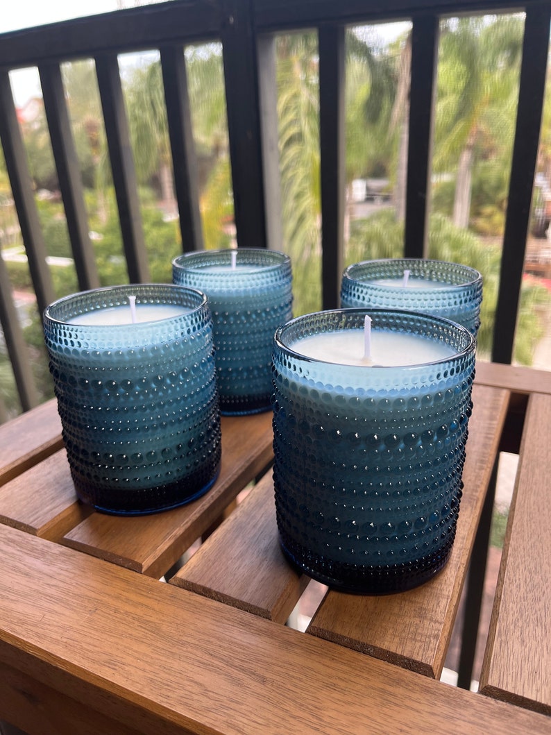 May include: Four blue glass candles with white wax and white wicks. The candles are sitting on a wooden table.