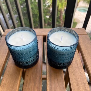 May include: Two blue glass candle jars with white wax and white wicks. The jars have a textured, bumpy surface.