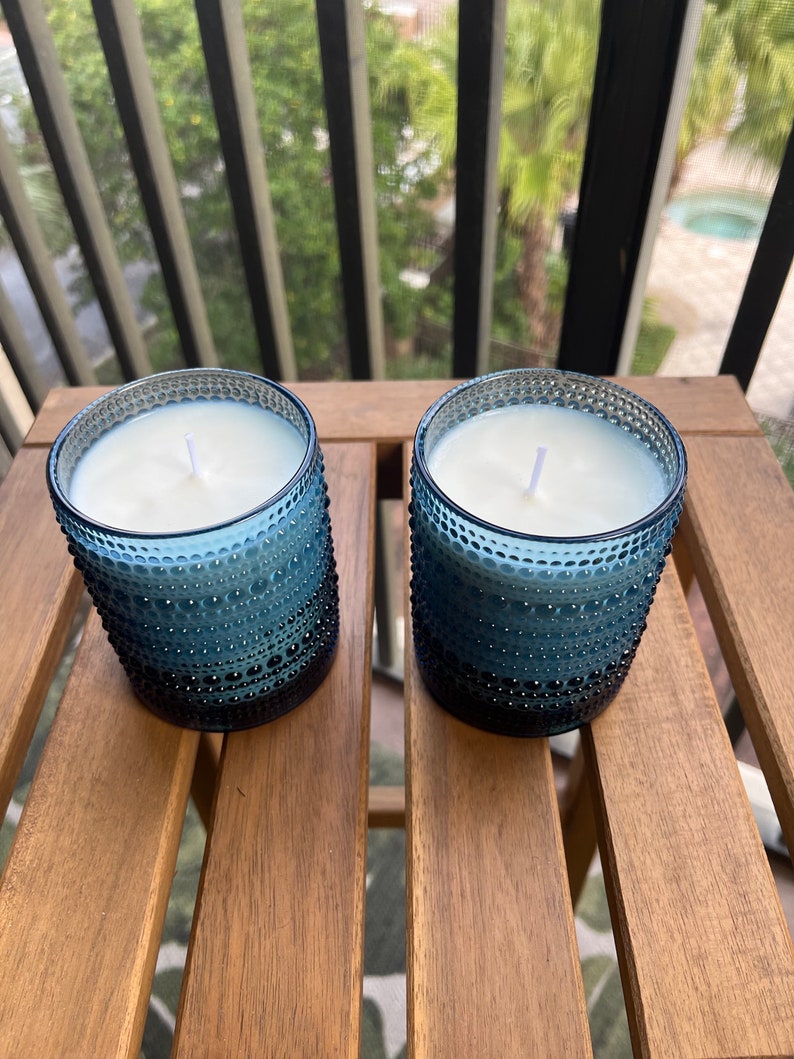 May include: Two blue glass candle jars with white wax and white wicks. The jars have a textured, bumpy surface.