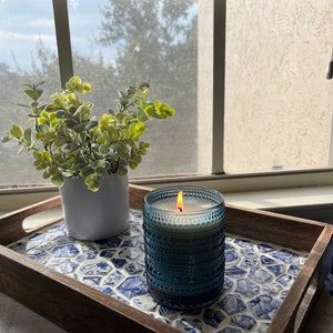 May include: A wooden tray with a blue and white mosaic tile design. A blue glass candle with a white candle inside is lit and sitting on the tray. A small potted plant is also on the tray.